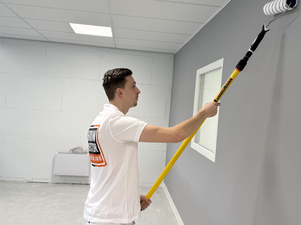 Schilder in wit poloshirt rolt een grijze wand naast een wit binnenraam onder een systeemplafond